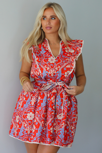 A woman modeling a sleeveless mini dress with a red, cream, and light blue paisley pattern, featuring a tie waist and ruffled collar, standing indoors against a white wall.