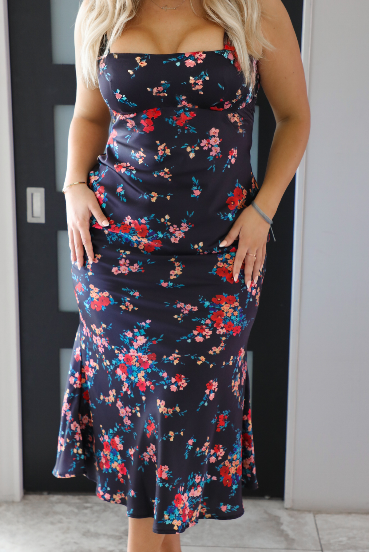 Close up of the midsection of a woman model wearing a navy dress with a multi-colored floral print. The dress is a satin material, is a midi length, and is curve-hugging. The photo is taken inside a modern white and black interior. 