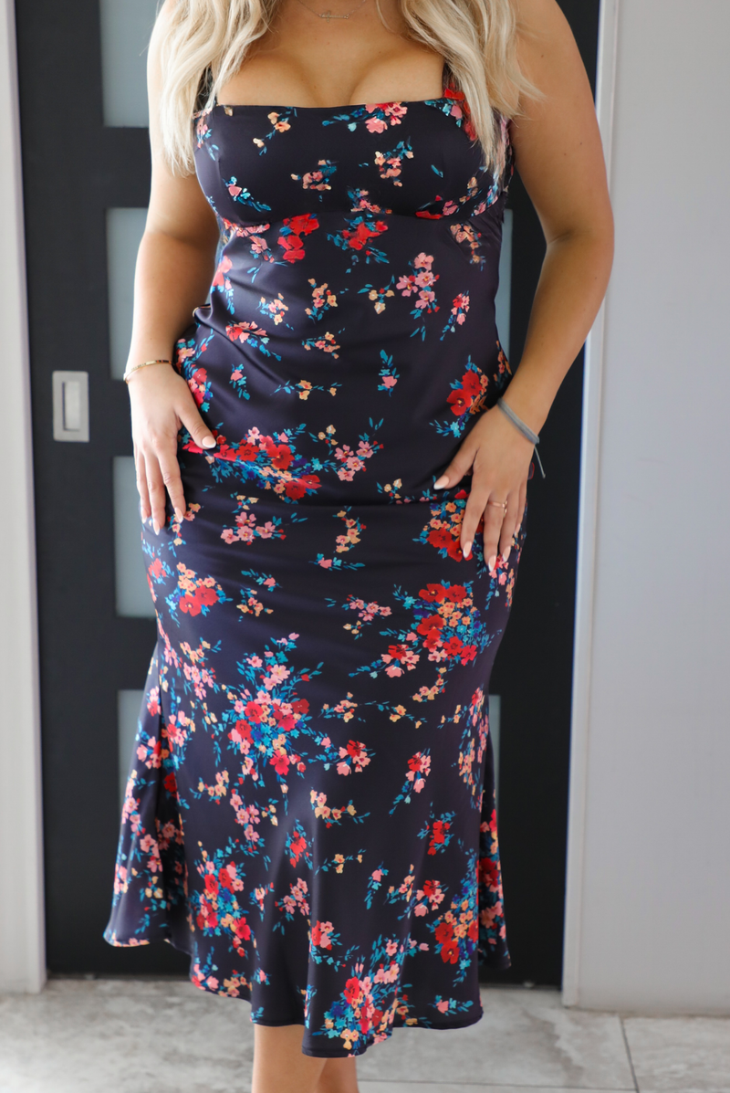 Close up of the midsection of a woman model wearing a navy dress with a multi-colored floral print. The dress is a satin material, is a midi length, and is curve-hugging. The photo is taken inside a modern white and black interior. 
