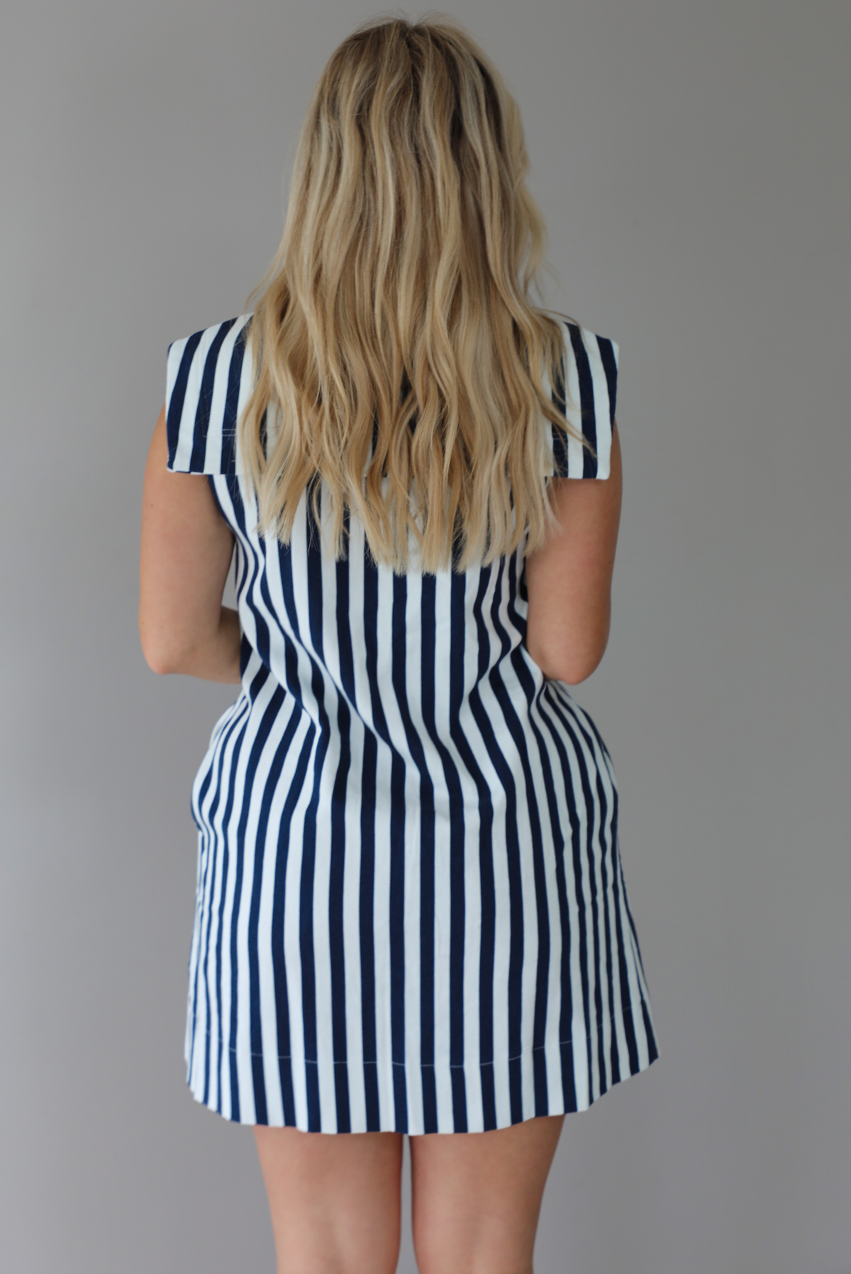 Woman wearing a navy and white striped dress with a bow detail at the neckline indoors against a white wall. The photo shows a closeup of the back of the dress. 