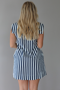 Woman wearing a navy and white striped dress with a bow detail at the neckline indoors against a white wall. The photo shows a closeup of the back of the dress. 