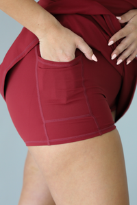 Woman wearing a maroon athleisure dress with thin straps and a relaxed fit indoors against a white wall. The photo shows a closeup of the shorts underneath with the models hand in the side pocket. 