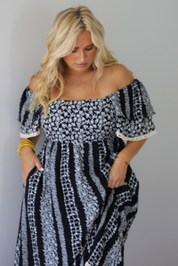A woman wearing a black and white maxi dress with a white floral print that alternates between a cluster appearance and a striped appearance, with off the shoulder sleeves and a flowy skirt, standing indoors against a white wall. Th photo shows closeup of the bodice and waist of the dress, showing it has pockets on either side. 