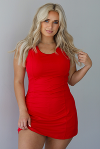 Woman wearing a red athleisure dress with a rounded neckline, thicker material, and a mini length indoors against a white wall.