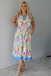 A woman wearing a short sleeved midi dress with a colorful tropical leaf print and light blue trim indoors against a white wall with white marble flooring. 