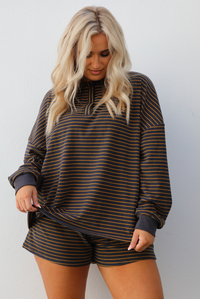 Close up of a woman model wearing a brown and multi-colored striped pattern throughout. The set consists of an oversized top with quarter-button details and shorts. The photo is taken outdoors against a white wall. The photo showcases the full length of the top that stops mid-hip on our 5'7" model.