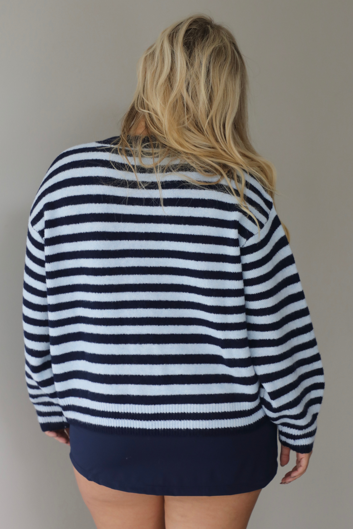 A woman shown from behind wearing a navy and blue striped cardigan paired with a navy two piece set underneath standing indoors against a white wall.