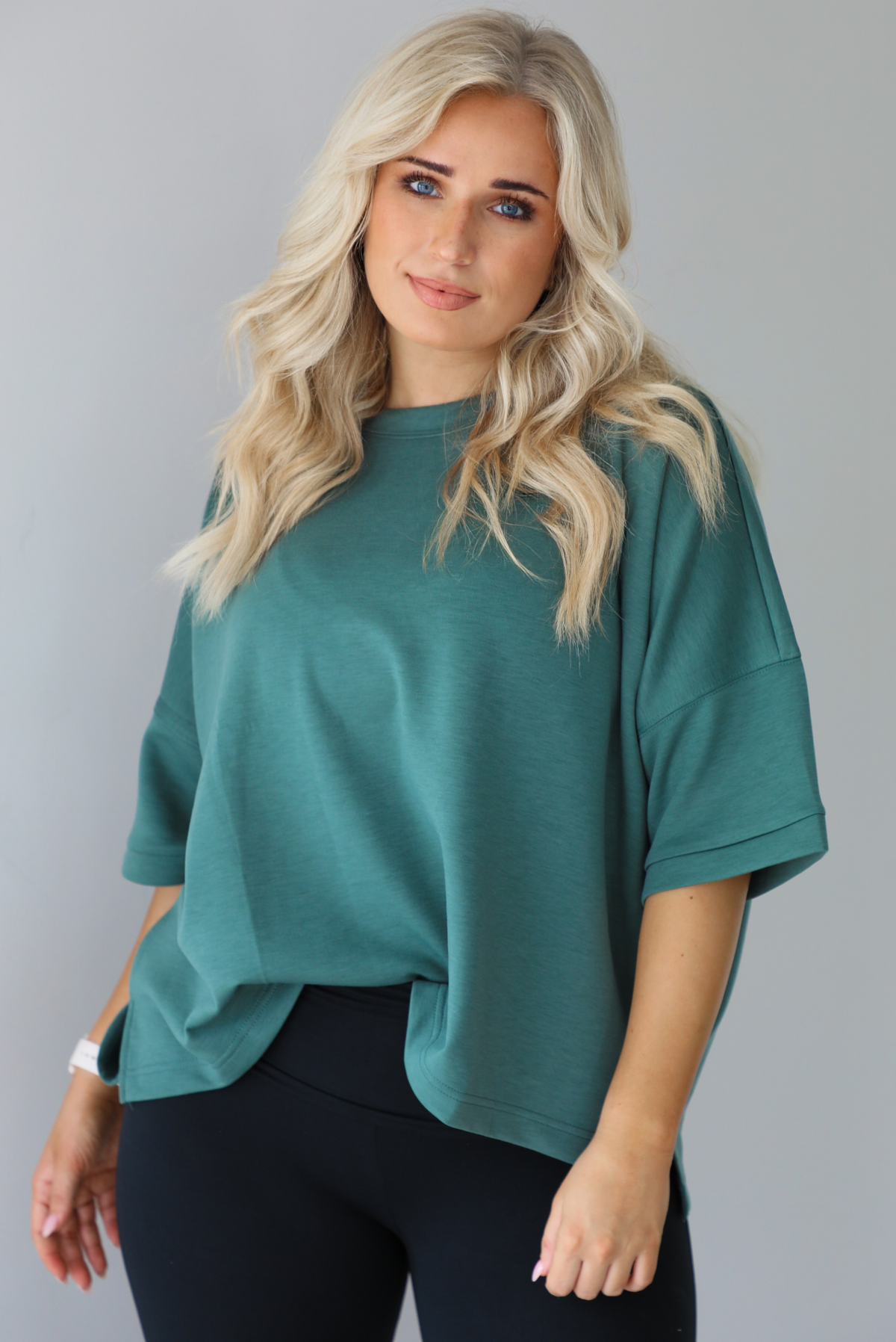 Woman in a teal oversized t-shirt paired with black leggings. The photo shows a closeup of the top which is tucked into the leggings in the front.The photo is taken indoors against a white wall.