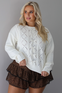 A woman wearing a white sweater with bow details along the front, untucked to show the full length, paired with black and brown leopard print frilly shorts with black lace trim detailing, standing indoors against a white wall.