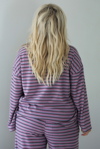 A woman shown from behind wearing a purple matching set with a black and blue striped pattern featuring an oversized long sleeved top and high waisted flared hem pants, standing indoors against a white wall. The photo shows a closeup of the back of the top.