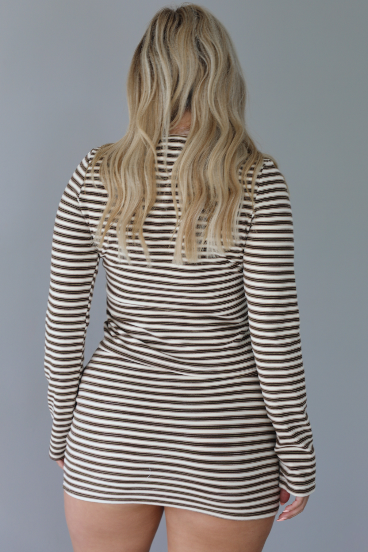 Woman wearing a brown and cream striped set featuring a button-down long sleeved top and mini skirt indoors against a white wall and shows the back of the set. 