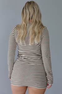 Woman wearing a brown and cream striped set featuring a button-down long sleeved top and mini skirt indoors against a white wall and shows the back of the set. 