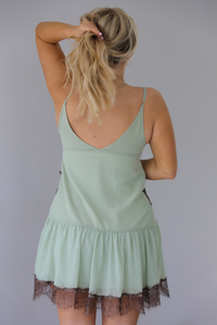 Woman wearing a sage green spaghetti strapped dress with brown lace detailing on the bodice and hem. The photo is taken indoors against a white wall and sows the back of the dress with its adjustable straps.