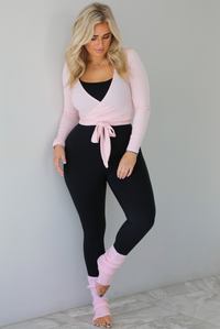 Woman wearing a pink cropped wrap style fitted top over a black fitted jumpsuit. The outfit is paired with pink leg warmers. The photo is taken indoors against a white wall with white marble flooring. 