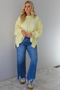 Woman wearing a pale yellow oversized sweater with dark wash denim and sneakers. The photo is taken indoors against a white wall with white marble flooring. 