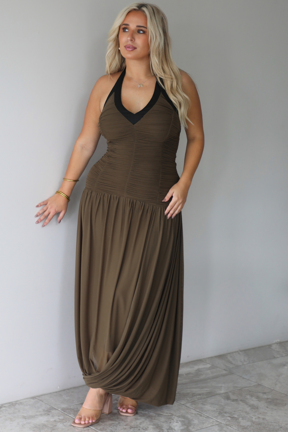 A woman wearing an olive maxi dress with black trim detailing at the v-halter neckline, has a fitted bodice and drop waist design, with a maxi-length flowy skirt, standing indoors against a white wall with white marble flooring. 