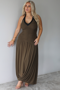 A woman wearing an olive maxi dress with black trim detailing at the v-halter neckline, has a fitted bodice and drop waist design, with a maxi-length flowy skirt, standing indoors against a white wall with white marble flooring. 