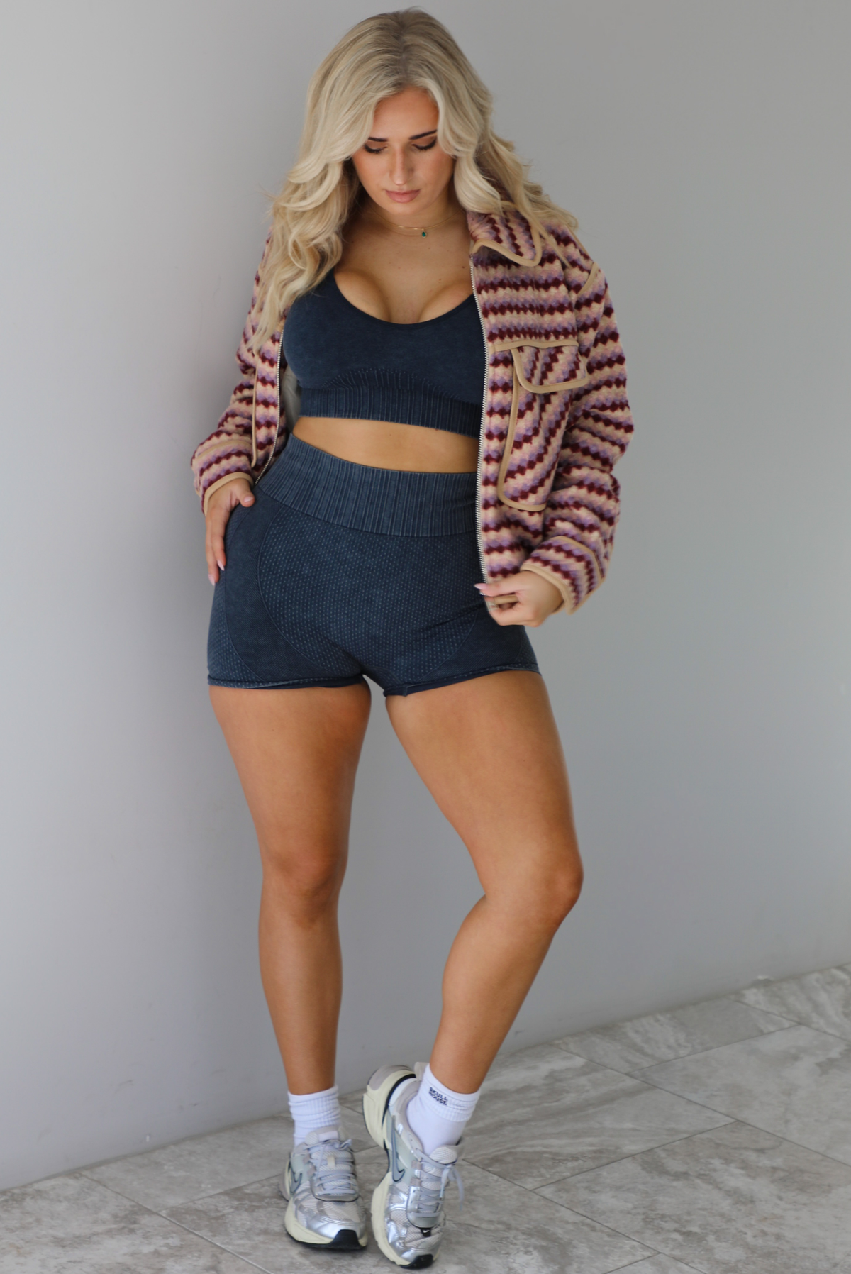 Woman wearing a pink, burgundy, lilac, and sand colored jacket with a charcoal colored two-piece set featuring a cropped sports bra and high waisted short biker shorts. The photo is taken indoors against a white wall with white marble flooring. 