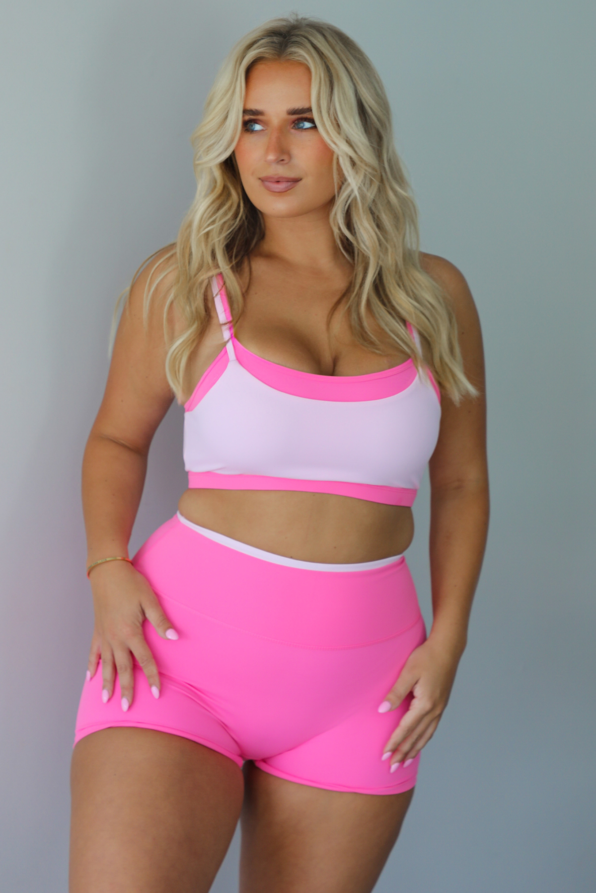 A woman modeling a bright pink and light pink two-piece activewear set consisting of a sports bra and matching high-waisted shorts, standing indoors against a white wall.