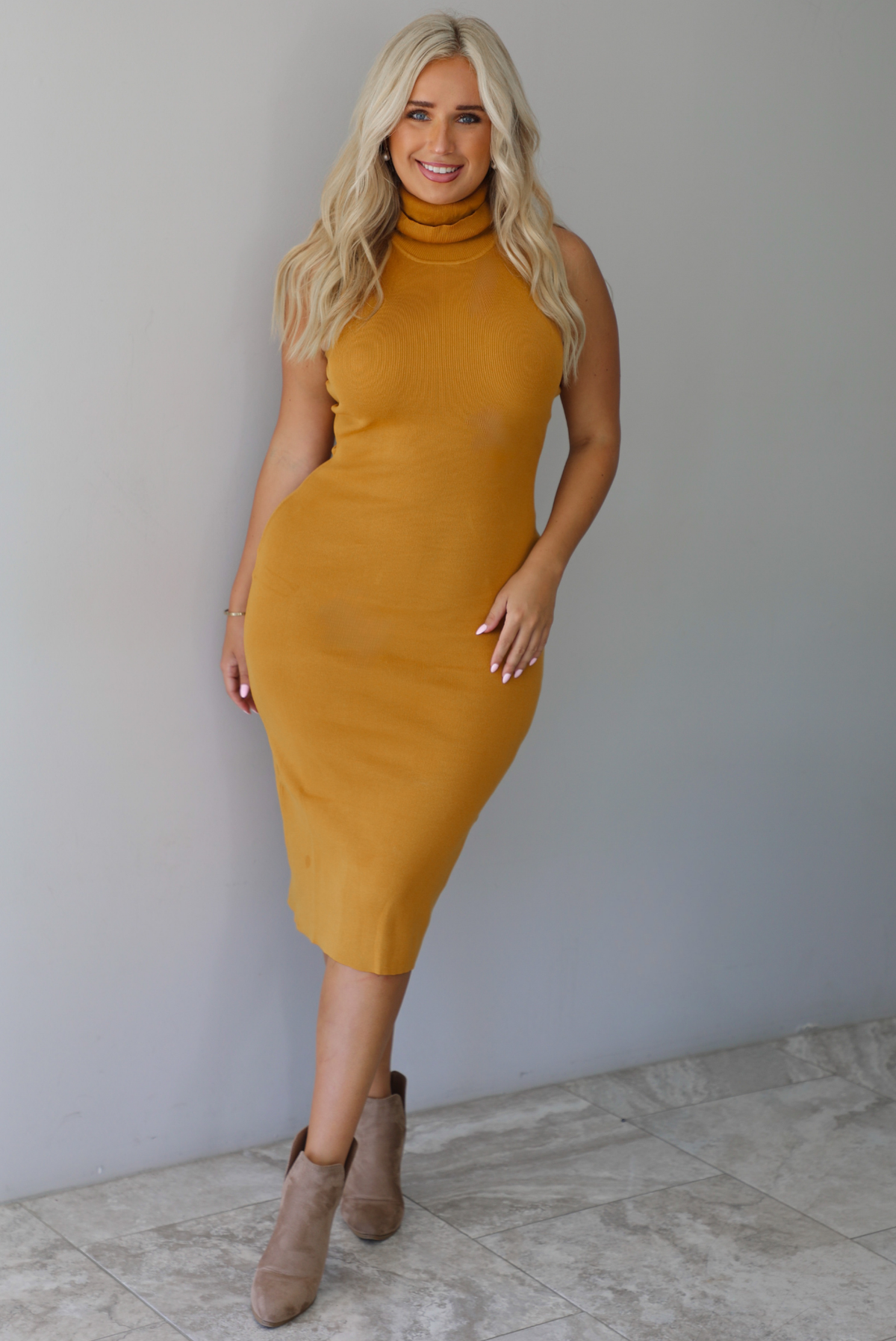 A woman wearing a marigold sleeveless midi dress with a high, turtle neckline, ribbed textured material, and is fitted, standing indoors against a white wall with white marble flooring. 