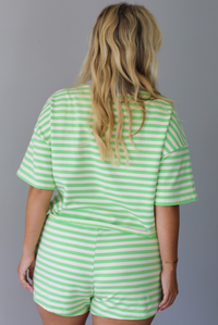 A woman modeling a matching green and white striped lounge set consisting of a short-sleeve top and shorts, with a light blue inner collar on the top and drawstring detail, standing indoors against a white wall. The photo shows the back of the set.