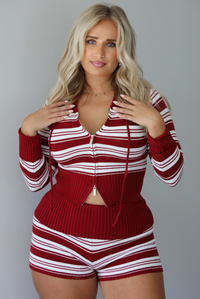 A woman modeling a matching red and white striped knit set with a front zipper, drawstring hood, paired with brown platform sandals, standing indoors against a white wall.