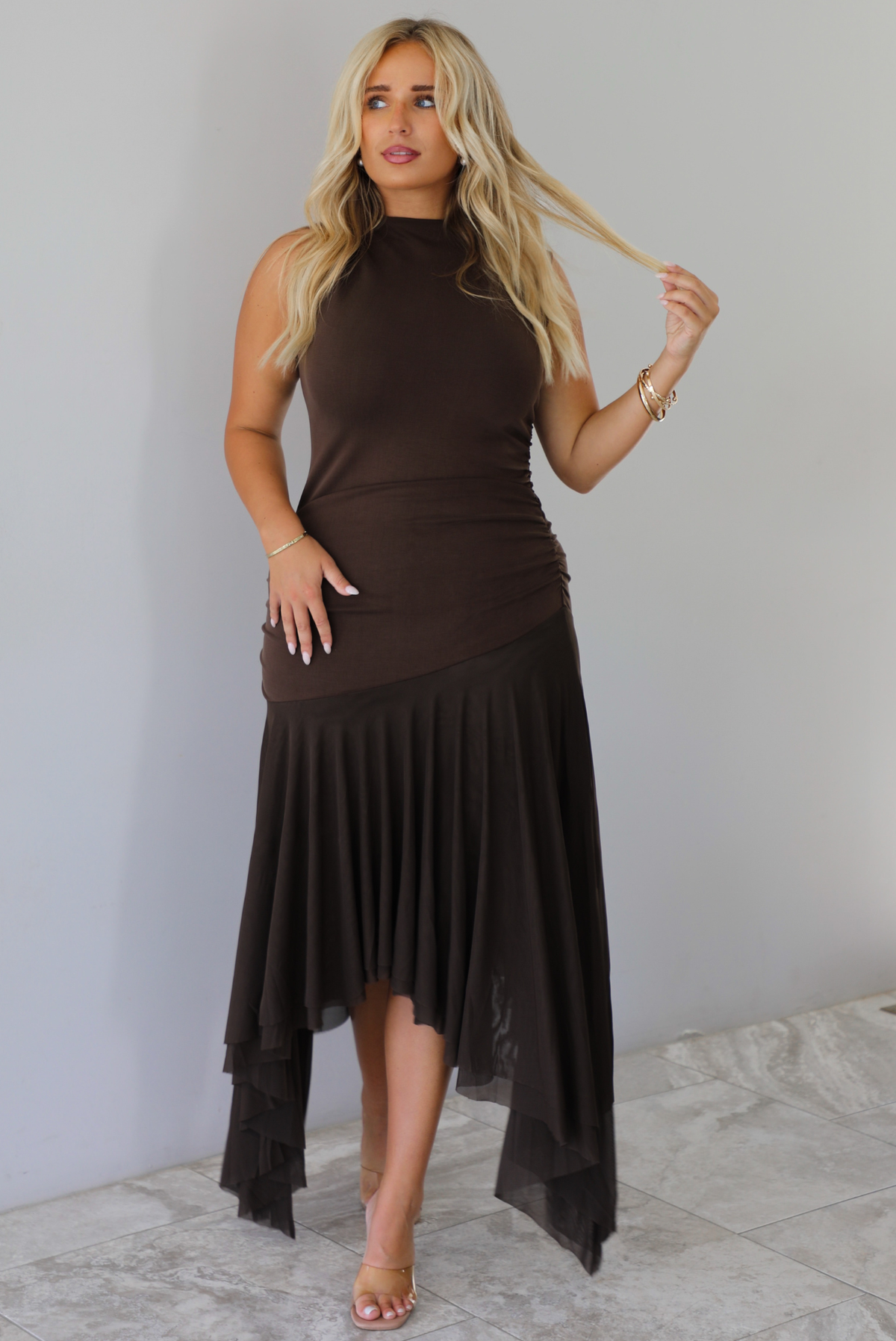 A woman wearing a brown midi dress that has a high neckline, is fitted until the upper thigh, then leads to sheer mesh that's flowy ands an asymmetrical hem, standing indoors against a white wall with white marble flooring. 