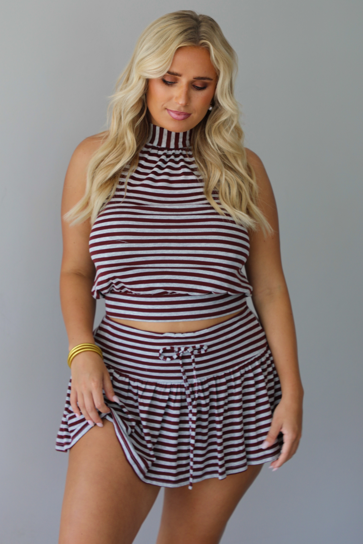A woman wearing a burgundy and grey striped two piece set featuring a high-neck cropped top and mini skirt, high scrunched white socks, and brown leather loafers, standing indoors against a white wall.