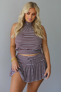 A woman wearing a burgundy and grey striped two piece set featuring a high-neck cropped top and mini skirt, high scrunched white socks, and brown leather loafers, standing indoors against a white wall.