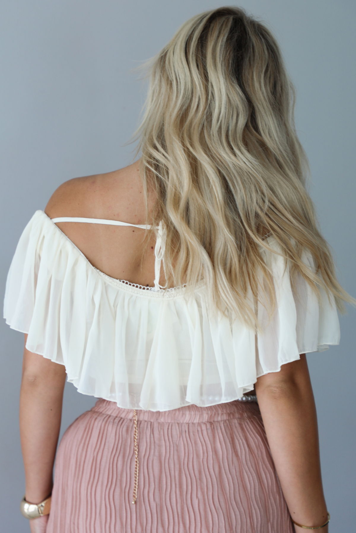 Woman wearing a cream cropped, off the shoulder top with a high waisted blush maxi skirt indoors against a white wall. The photo shows the back of the top. 