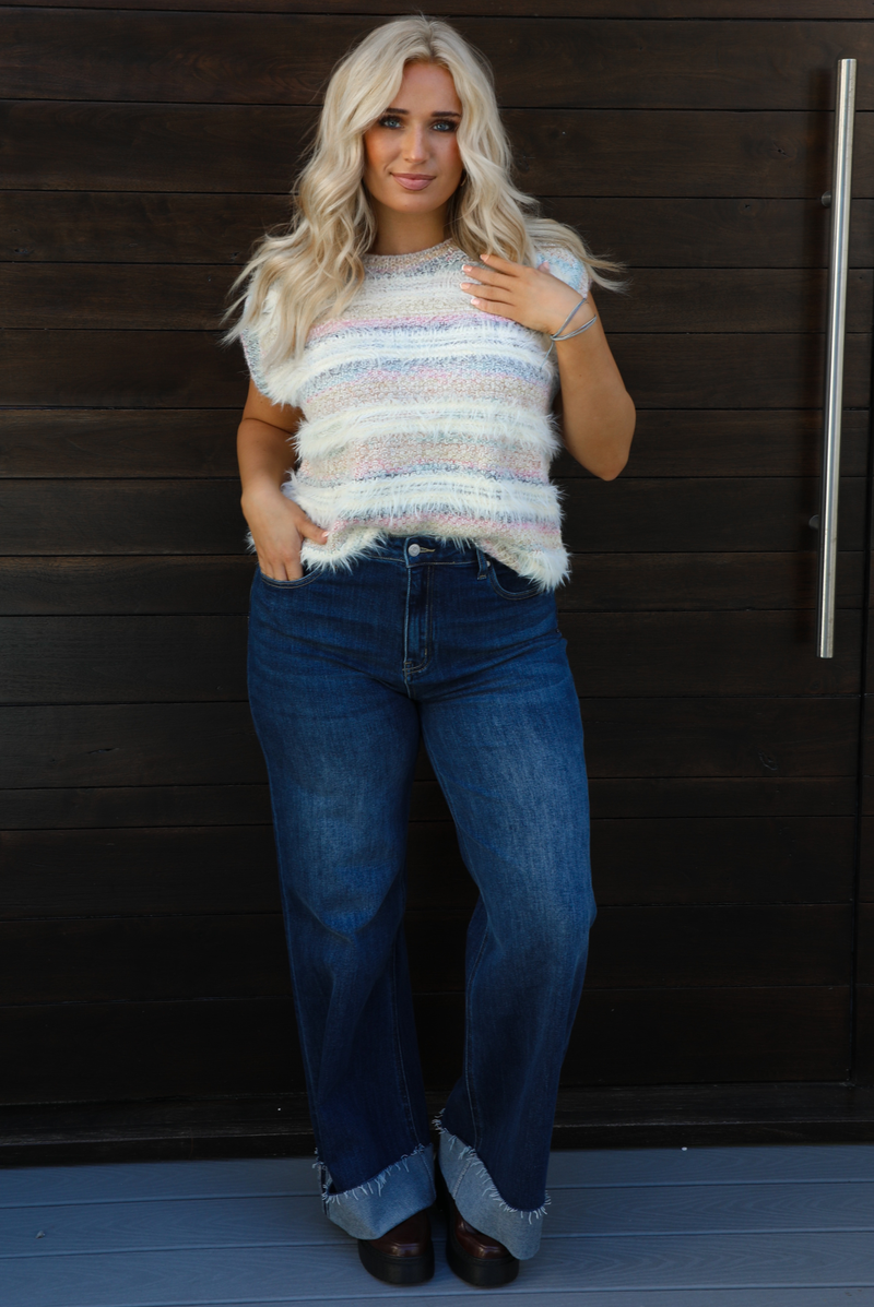 Woman model wearing a soft, striped sweater vest with dark wash denim jeans. The photo is taken outdoors against a dark brown wooden door. 
