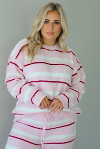 Woman wearing a pink set with a darker pink and white striped design. The set consists of an oversized sweater top and high waisted pants. The sweater of the set is tucked slightly in the front of the pants. The photo shows a closeup of the sweater. The photo is taken indoors against a white wall. 