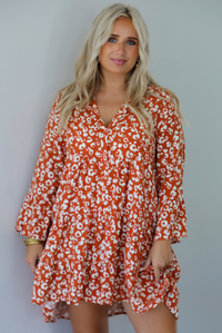 A woman wearing an orange and white animal print long sleeved, flowy mini dress, standing indoors against a white wall.