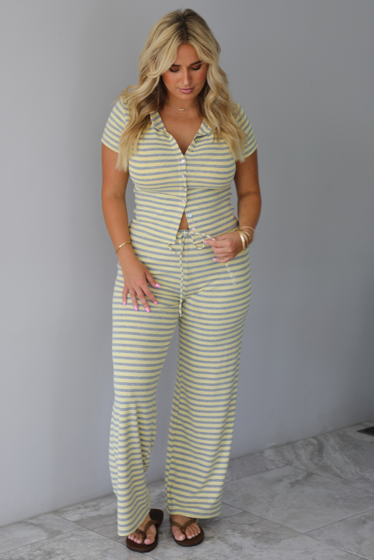 A woman wearing a two piece set consisting of a collared, button-down, short sleeve top and high waisted, drawstring, wide leg pants, standing indoors against a white wall with white marble flooring. 