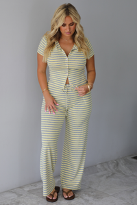 A woman wearing a two piece set consisting of a collared, button-down, short sleeve top and high waisted, drawstring, wide leg pants, standing indoors against a white wall with white marble flooring. 