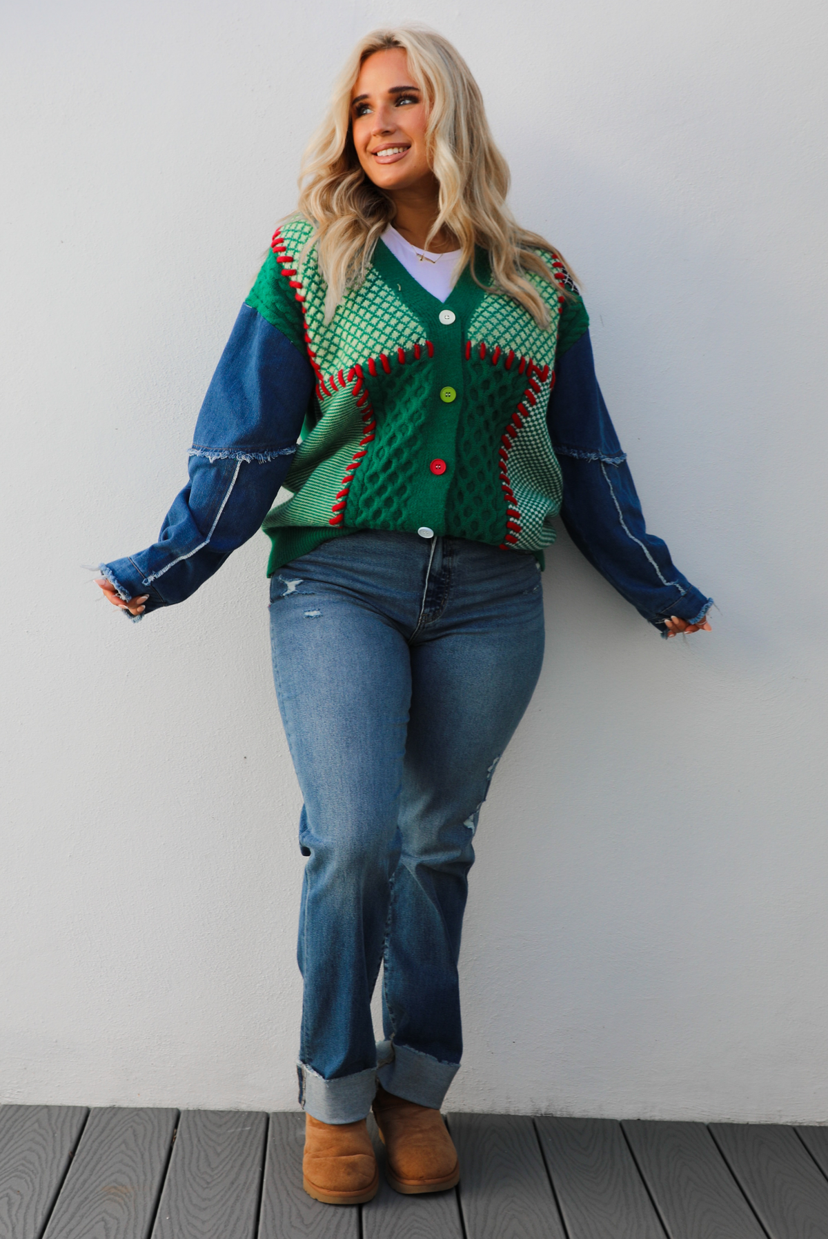 Woman model wearing a green and multi-colored cardigan with denim sleeves, medium wash denim jeans, and tan uggs. The photo is taken against a white wall outdoors.