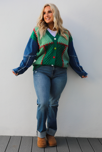 Woman model wearing a green and multi-colored cardigan with denim sleeves, medium wash denim jeans, and tan uggs. The photo is taken against a white wall outdoors.