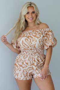 Woman wearing a tan and white romper that has off the shoulder short puffed sleeves, a cinched waistline, and a repeating leaf pattern indoors against a white wall.
