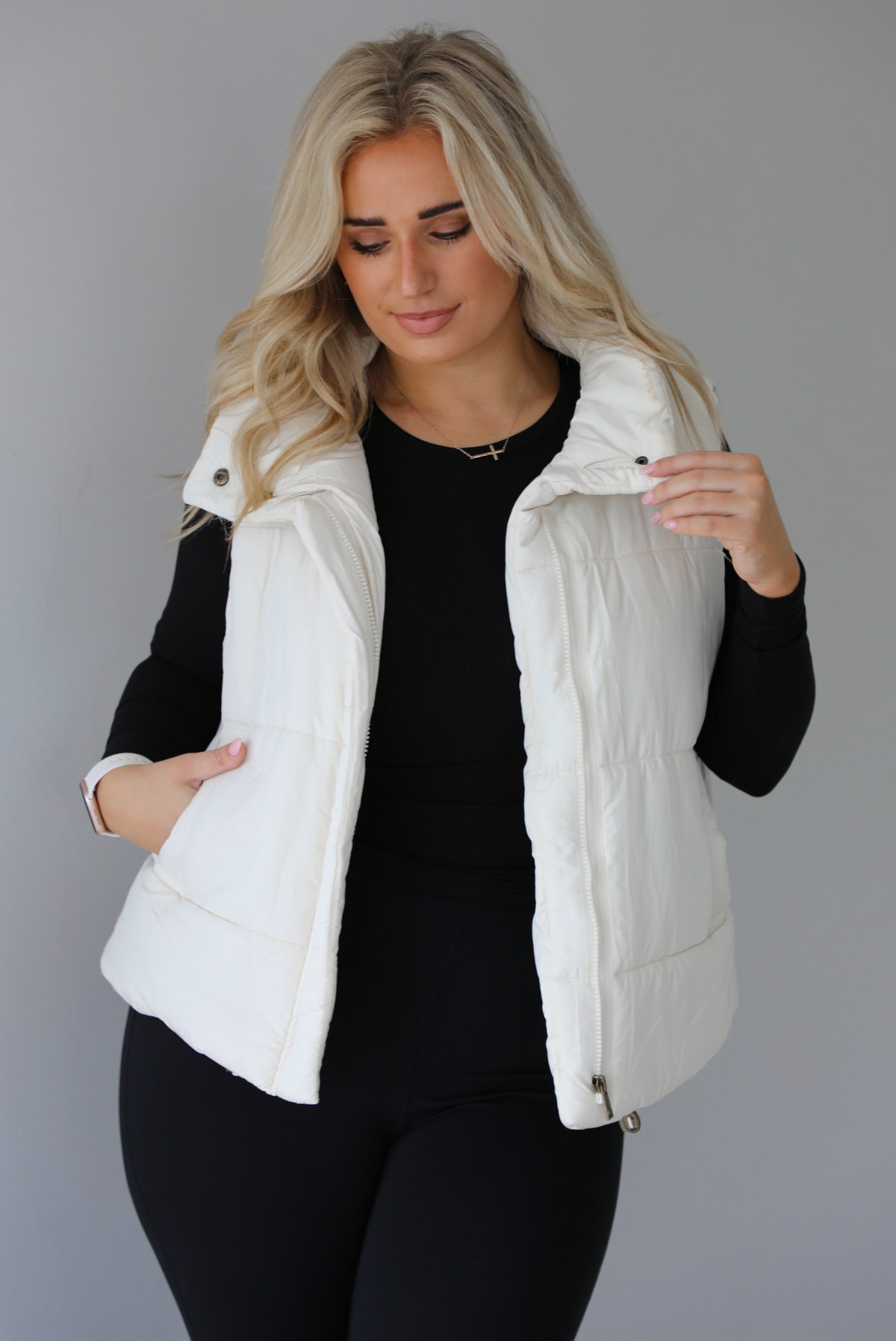 Woman wearing a light sand colored puffer vest with a fitted black long sleeved top and fitted leggings. The vest has functional button closures at the top, a zipper closure in the front, and functional pockets on either side. The photo shows the vest unzipped. The photo is taken indoors against a white wall with white marble flooring. 