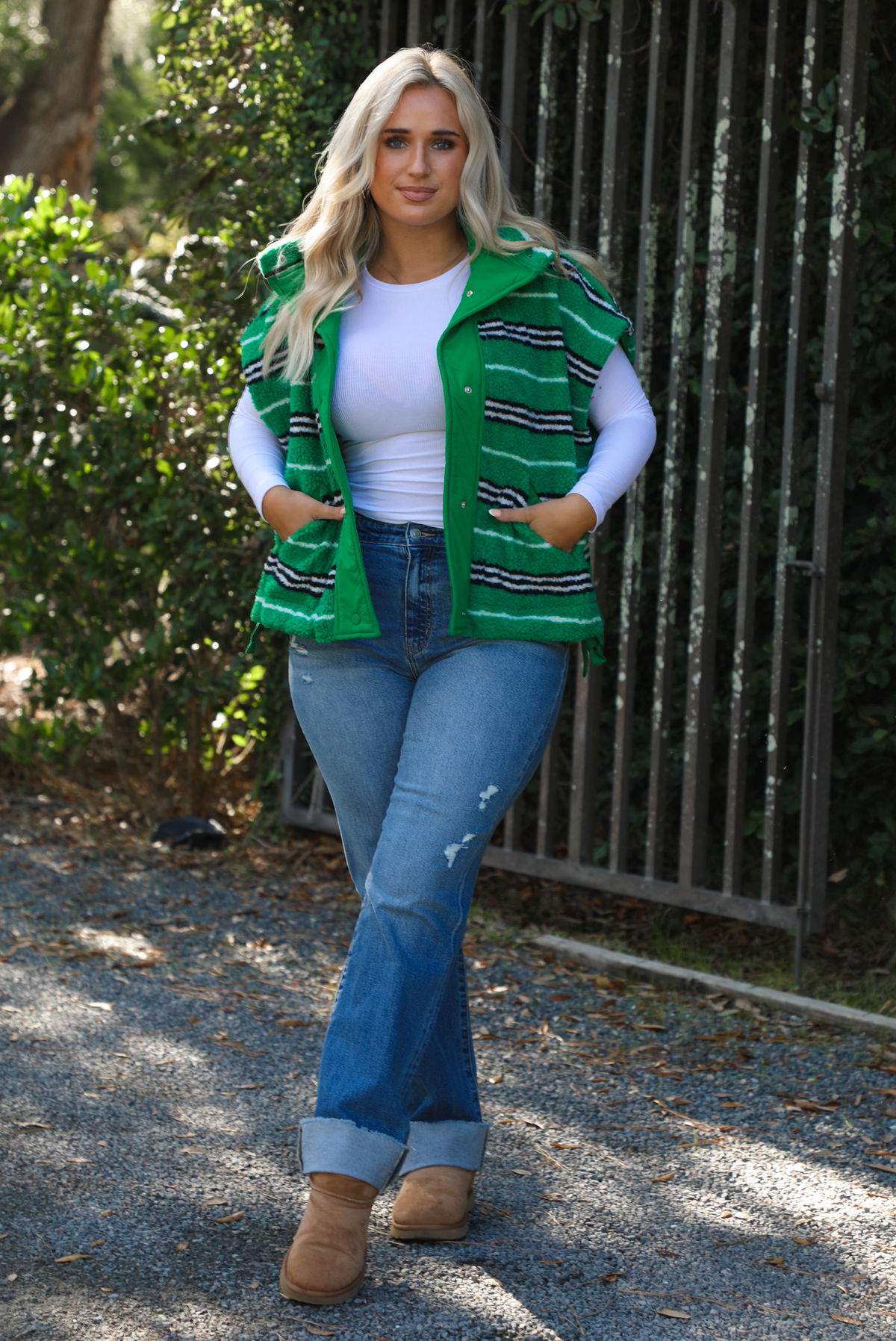 Woman model wearing a green, black, and white striped vest that has a sherpa texture to it, functional button closures in the front, and tie closures on either side, It is paired with a white long sleeved top, a medium wash pair of denim jeans, and tan ugg boots. The photo is taken outdoors with a metal gate and greenery in the back. The vest is shown unbuttoned.