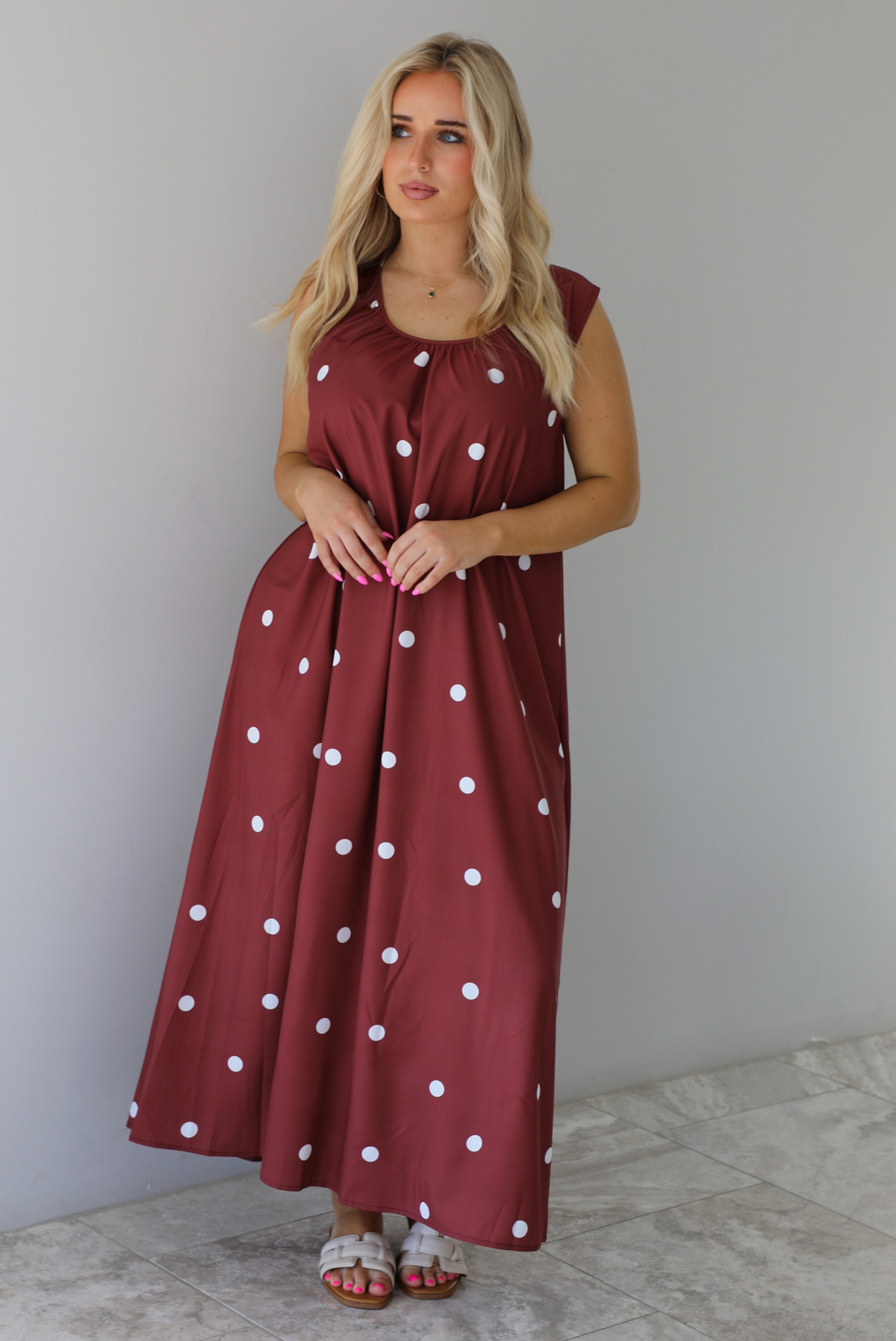 Woman wearing a red brown maxi length dress that has a white polka dot print pattern throughout. The dress is paired with sand sandals. The photo is taken indoors against a white wall with white marble flooring. 