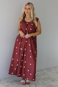 Woman wearing a red brown maxi length dress that has a white polka dot print pattern throughout. The dress is paired with sand sandals. The photo is taken indoors against a white wall with white marble flooring. 