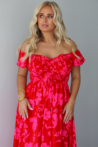 A closeup of a woman modeling a bright red off-the-shoulder maxi dress with a vibrant pink floral pattern, focusing in on the smocked bodice and drawstring detail in the center of the bodice, standing indoors agains a white wall.