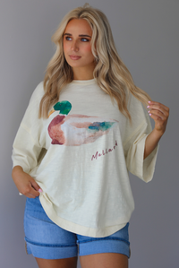 Woman wearing an off-white t-shirt with a graphic design of a mallard duck on the front with blue denim shorts. The photo is taken indoors against a white wall.