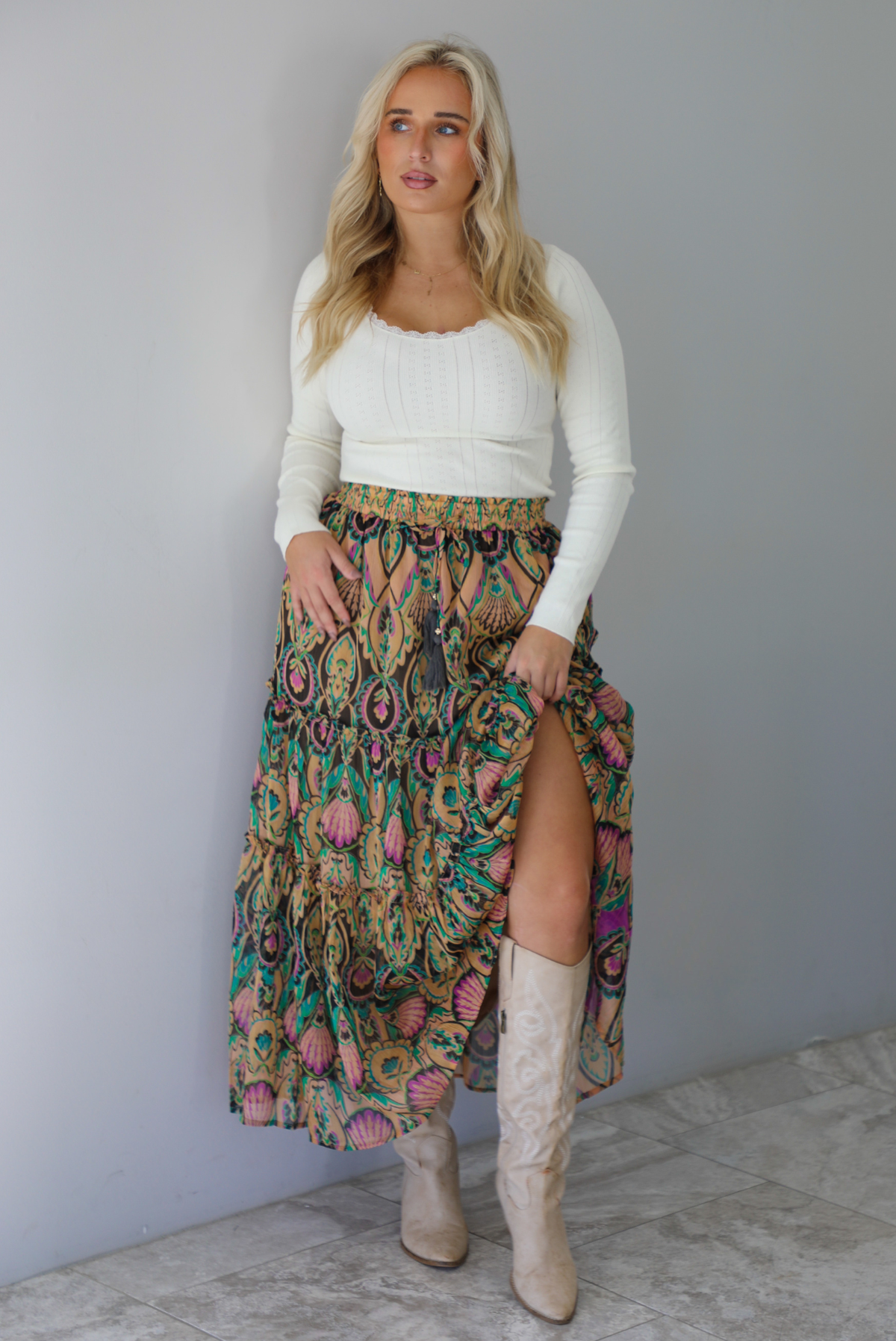 A woman wearing a long sleeved cream top and multi-colored maxi length skirt paired with knee high sand colored ankle boots, standing indoors against a white wall with white marble flooring. 