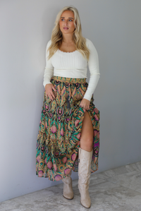 A woman wearing a long sleeved cream top and multi-colored maxi length skirt paired with knee high sand colored ankle boots, standing indoors against a white wall with white marble flooring. 