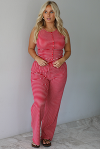 A woman modeling a red and white striped sleeveless set consisting of a tank top with non-functional button closures in the front and high waisted pants with a drawstring waistline and pockets on either side indoors against a white wall with white marble flooring. 