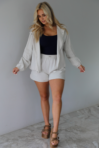 Woman wearing a stone colored two piece set featuring a zip up jacket and shorts with a navy tank tucked into the shorts indoors against a white wall with white marble flooring. 