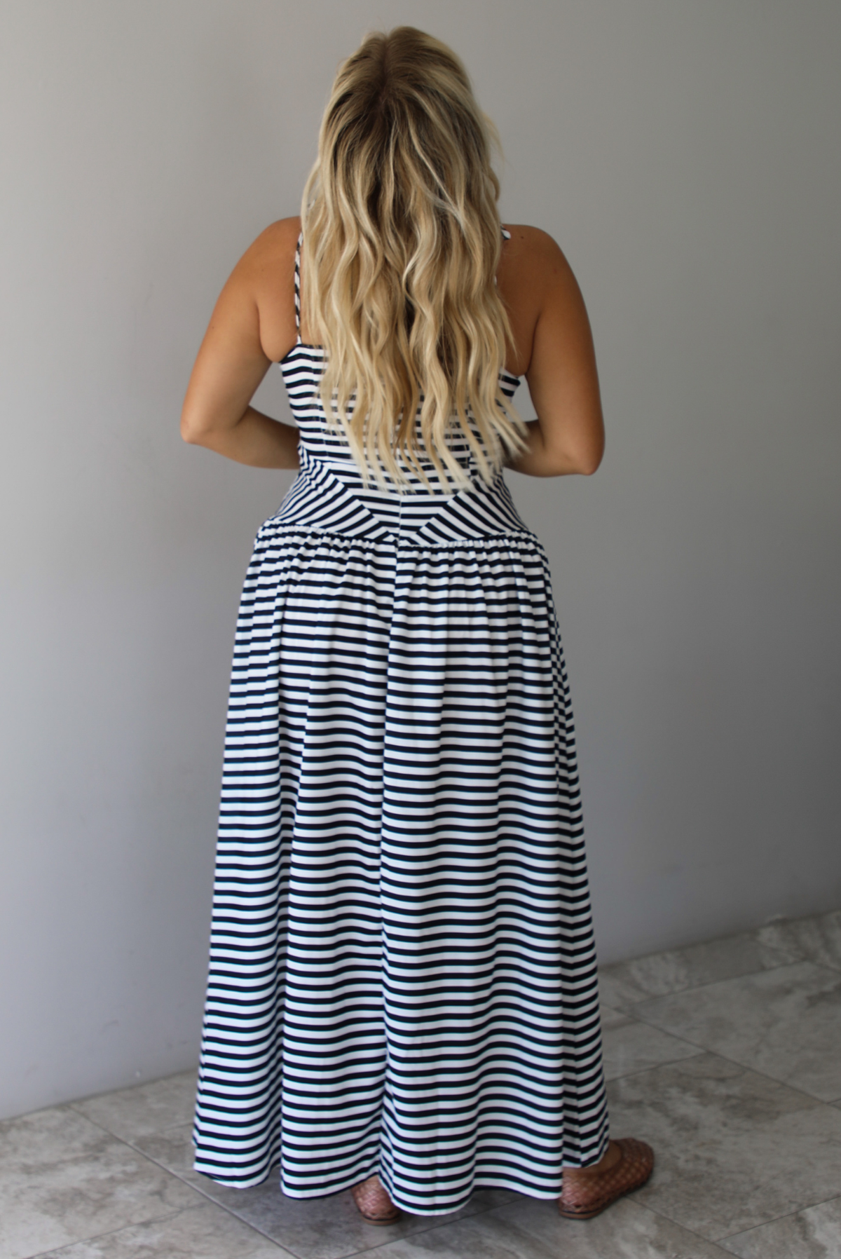 A woman viewed from behind wearing a sleeveless black and white horizontally striped maxi dress, standing indoors against a white wall with white marble flooring. 