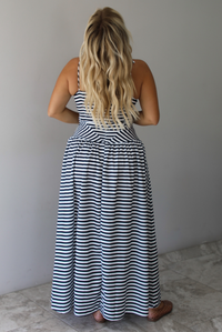 A woman viewed from behind wearing a sleeveless black and white horizontally striped maxi dress, standing indoors against a white wall with white marble flooring. 